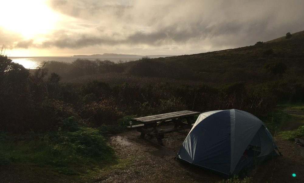A blue tent pitched near the Pacific coast with the sun setting.