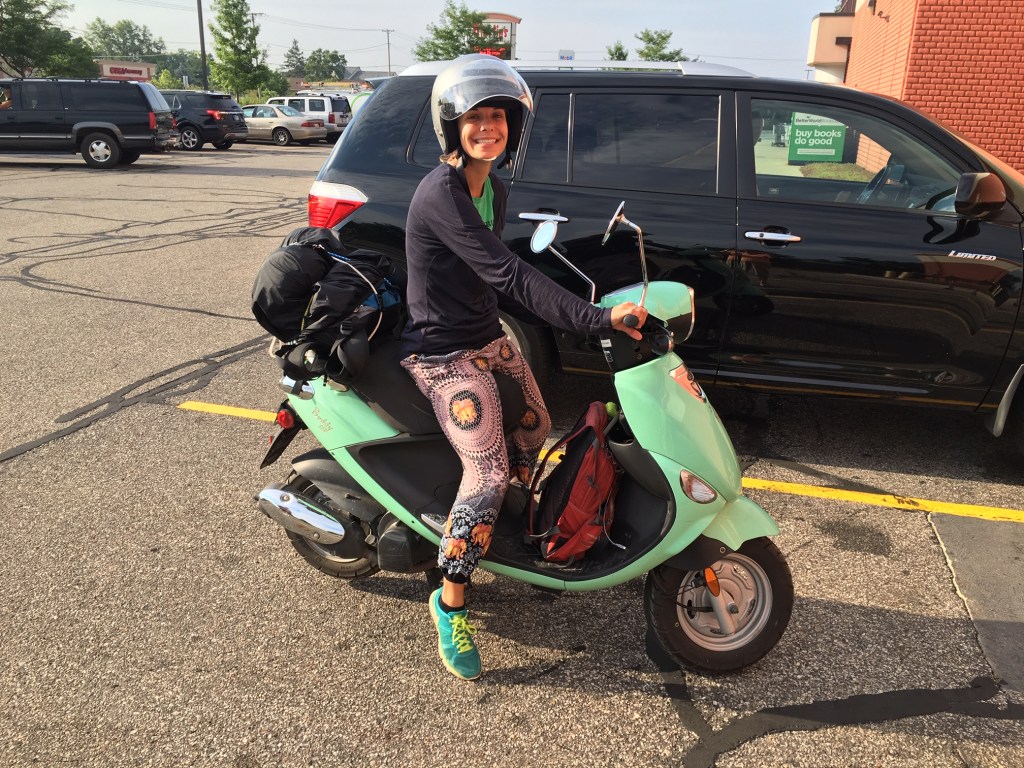 A smiling woman on a parked teal moped.